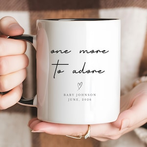 May include: White ceramic mug with black interior and handle. The mug features the text "one more to adore" in a handwritten font, along with a heart symbol and the name "Baby Johnson" and the date "June, 2026".