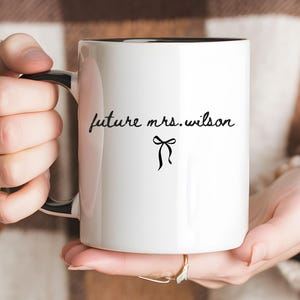 May include: A white ceramic mug with a black interior and handle. The exterior features the black text "future mrs. wilson" in a cursive script font, positioned above a small, simple black line drawing of a decorative bow centered on the mug.