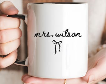 May include: A white ceramic mug with a black interior and handle. The exterior features the black text "mrs. wilson" in a cursive script font, positioned above a small, simple black line drawing of a decorative bow centered on the mug.