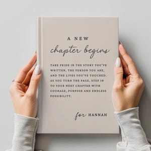 May include: A light beige hardcover book featuring the black text A NEW chapter begins followed by a paragraph about turning the page and stepping into a new chapter with courage, purpose, and endless possibility, ending with for HANNAH.
