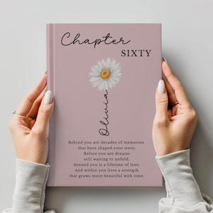 May include: A dusty rose hardcover book featuring the text Chapter SIXTY, a white daisy illustration, the name Olivia in script, and a printed poem that reads: Behind you are decades of memories that have shaped your story, Before you are dreams still waiting to unfold, Around you is a lifetime of love, And within you lives a strength that grows more beautiful with time.