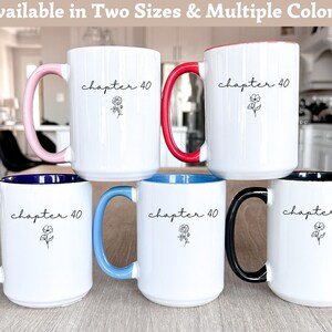 May include: Five white ceramic mugs with the text "chapter 40" and a small floral line drawing. Each mug features a different colored handle and interior rim: light pink, red, dark blue, light blue, and black. The mugs are arranged in a group.