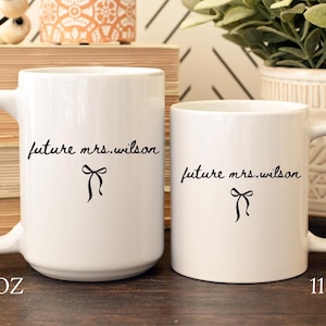 K&ouml;nnte beinhalten: Zwei wei&szlig;e Keramikbecher mit schwarzem Text "future mrs. wilson" und einer Schleifengrafik. Der gr&ouml;&szlig;ere Becher ist mit "15 OZ" und der kleinere Becher mit "11 OZ" beschriftet.