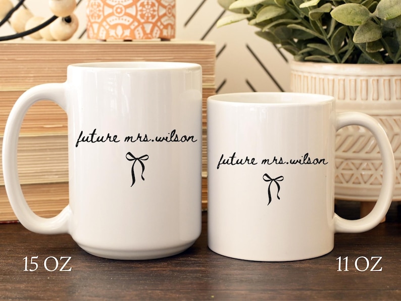 K&ouml;nnte beinhalten: Zwei wei&szlig;e Keramikbecher mit schwarzem Text "future mrs. wilson" und einer Schleifengrafik. Der gr&ouml;&szlig;ere Becher ist mit "15 OZ" und der kleinere Becher mit "11 OZ" beschriftet.