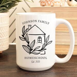 May include: White ceramic mug with a black illustration of a house, leaves, and text. The text reads "JOHNSON FAMILY HOMESCHOOL Est. 2025". The mug has a large handle and is on a wooden surface.