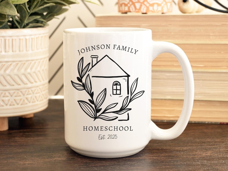 May include: White ceramic mug with a black illustration of a house, leaves, and text. The text reads "JOHNSON FAMILY HOMESCHOOL Est. 2025". The mug has a large handle and is on a wooden surface.