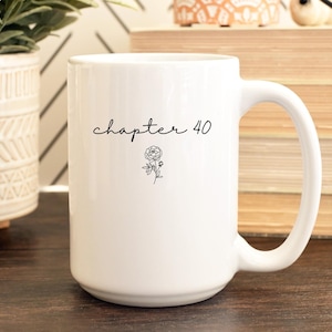 May include: A white ceramic mug featuring the black text "chapter 40" in a cursive script font, positioned above a small, detailed black line drawing of a single flower. The mug has a smooth, cylindrical body and a curved handle on the right side.