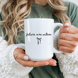 K&ouml;nnte beinhalten: Wei&szlig;er Keramikbecher mit dem Text "future mrs. wilson" in handschriftlichem Stil, mit einer schwarzen Schleifen-Grafik. Der Becher wird von einer Person gehalten, die ein gr&uuml;nes Hemd und einen wei&szlig;en Pullover tr&auml;gt.