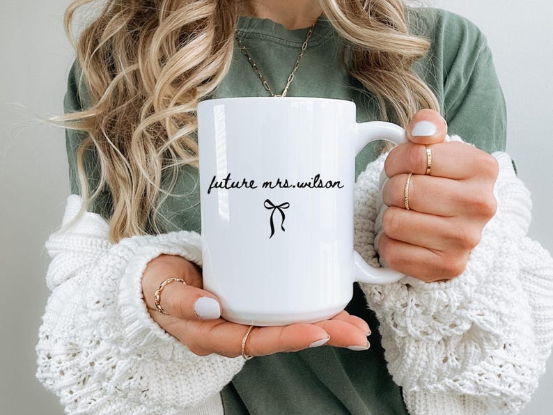 K&ouml;nnte beinhalten: Wei&szlig;er Keramikbecher mit dem Text "future mrs. wilson" in handschriftlichem Stil, mit einer schwarzen Schleifen-Grafik. Der Becher wird von einer Person gehalten, die ein gr&uuml;nes Hemd und einen wei&szlig;en Pullover tr&auml;gt.