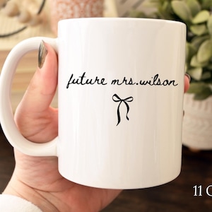 K&ouml;nnte beinhalten: Wei&szlig;e Keramiktasse mit dem Text "future mrs. wilson" in schwarzer Schrift und einer schwarzen Schleifengrafik. Die Tasse hat ein Fassungsverm&ouml;gen von 312 Gramm und einen gebogenen Griff.
