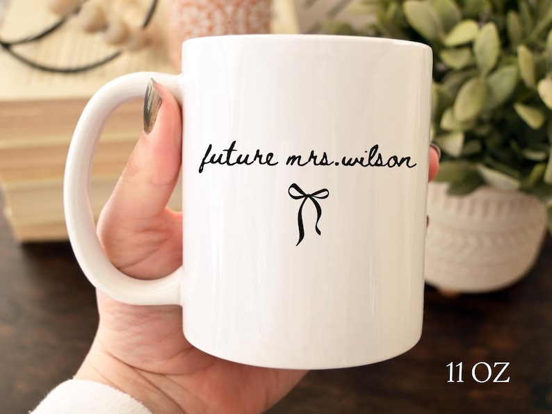 K&ouml;nnte beinhalten: Wei&szlig;e Keramiktasse mit dem Text "future mrs. wilson" in schwarzer Schrift und einer schwarzen Schleifengrafik. Die Tasse hat ein Fassungsverm&ouml;gen von 312 Gramm und einen gebogenen Griff.