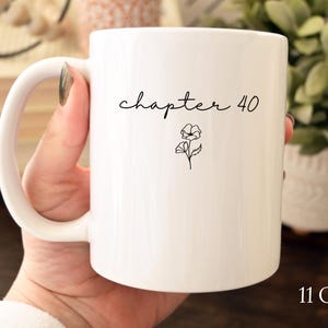 May include: A white ceramic mug with a curved handle, featuring the black text "chapter 40" in a cursive script font above a small, simple line drawing of a flower. The mug is labeled with the capacity of 11 oz in the bottom right corner.