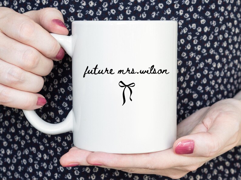 K&ouml;nnte beinhalten: Wei&szlig;e Keramik-Tasse mit dem Text "future mrs. wilson" in schwarzer Schrift, begleitet von einer schwarzen Schleifen-Illustration. Die Tasse wird von H&auml;nden mit rosa Nagellack vor einem dunklen floralen Hintergrund gehalten.