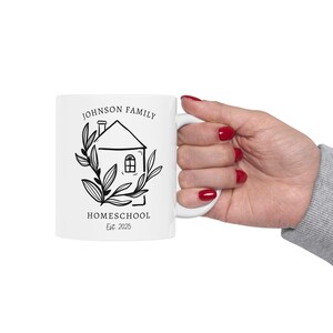 May include: White ceramic mug featuring a black and white illustration of a house with the words "JOHNSON FAMILY HOMESCHOOL Est. 2025". The design includes a house, leaves, and text. The mug is held by a hand with red nail polish.