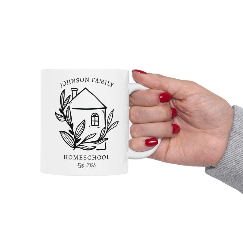 May include: White ceramic mug featuring a black and white illustration of a house with the words "JOHNSON FAMILY HOMESCHOOL Est. 2025". The design includes a house, leaves, and text. The mug is held by a hand with red nail polish.