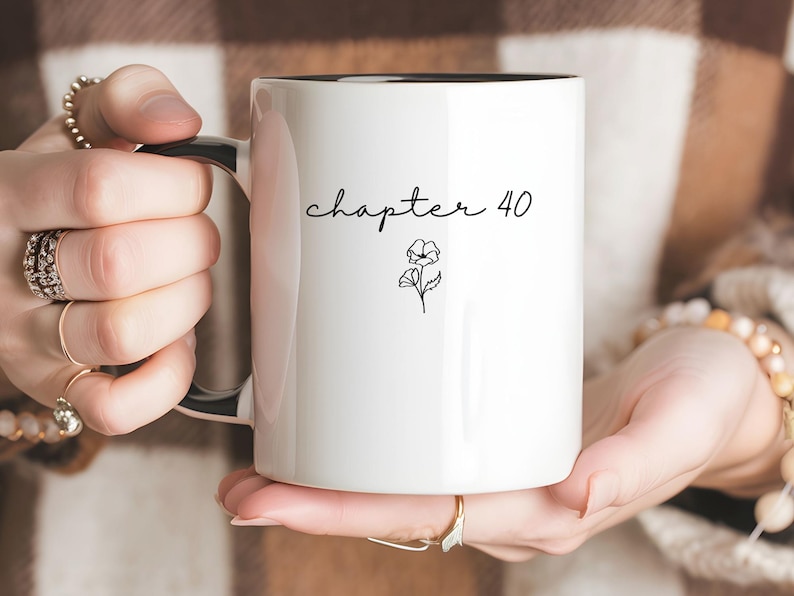 May include: A white ceramic mug with a black interior and handle. The exterior features the black text "chapter 40" in a cursive script, positioned above a small, simple black line drawing of a flower with two blossoms and leaves.