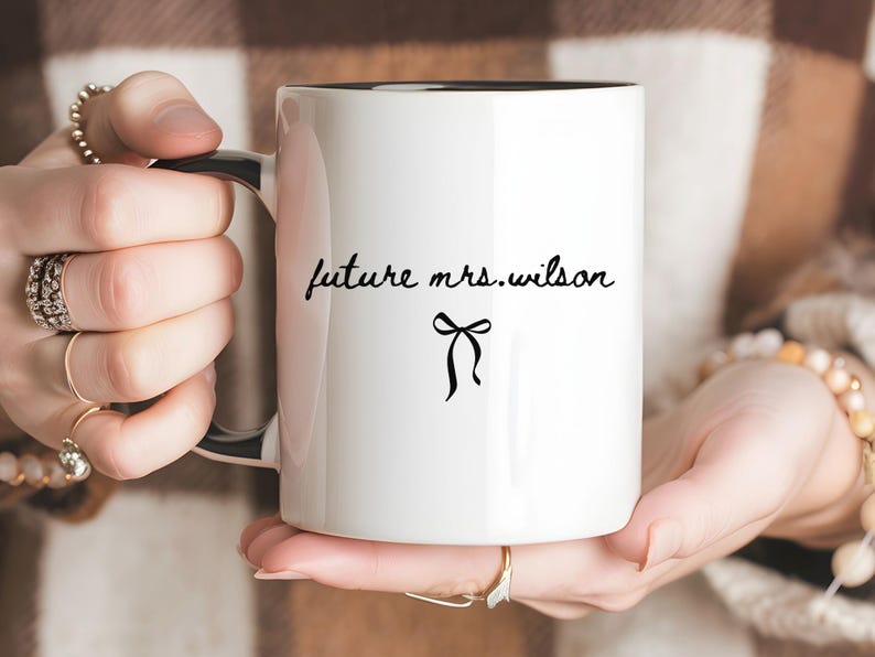 K&ouml;nnte beinhalten: Wei&szlig;er Keramikbecher mit schwarzem Innenraum und Henkel. Der Becher tr&auml;gt den Text "future mrs.wilson" in handschriftlichem Stil mit einer kleinen Schleifen-Grafik darunter. Der Becher wird von einer Person gehalten.