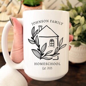 May include: White ceramic mug with a black line drawing of a house, leaves, and text that reads "JOHNSON FAMILY HOMESCHOOL Est. 2025". The mug is held by a person.