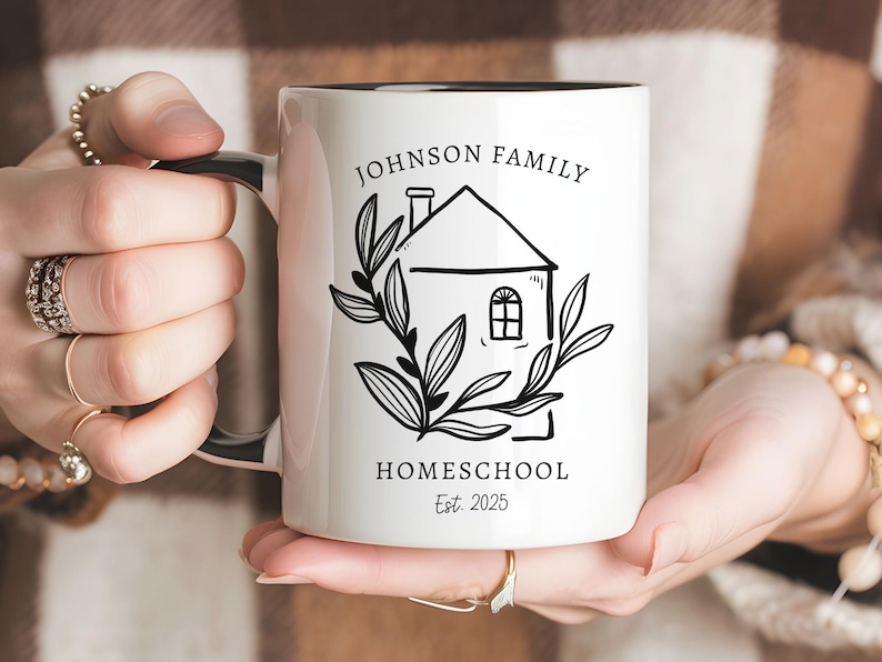 May include: White ceramic mug with a black interior and handle. The mug features a black and white illustration of a house with the text "JOHNSON FAMILY HOMESCHOOL Est. 2025" in a decorative font. The house is surrounded by a leafy wreath.