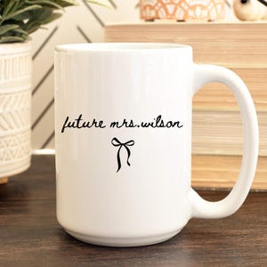 K&ouml;nnte beinhalten: Wei&szlig;er Keramikbecher mit gro&szlig;em Henkel. Der Becher tr&auml;gt den Text "future mrs. wilson" in handschriftlicher Form, mit einer schwarzen Schleifengrafik darunter. Der Becher steht auf einer Holzoberfl&auml;che.