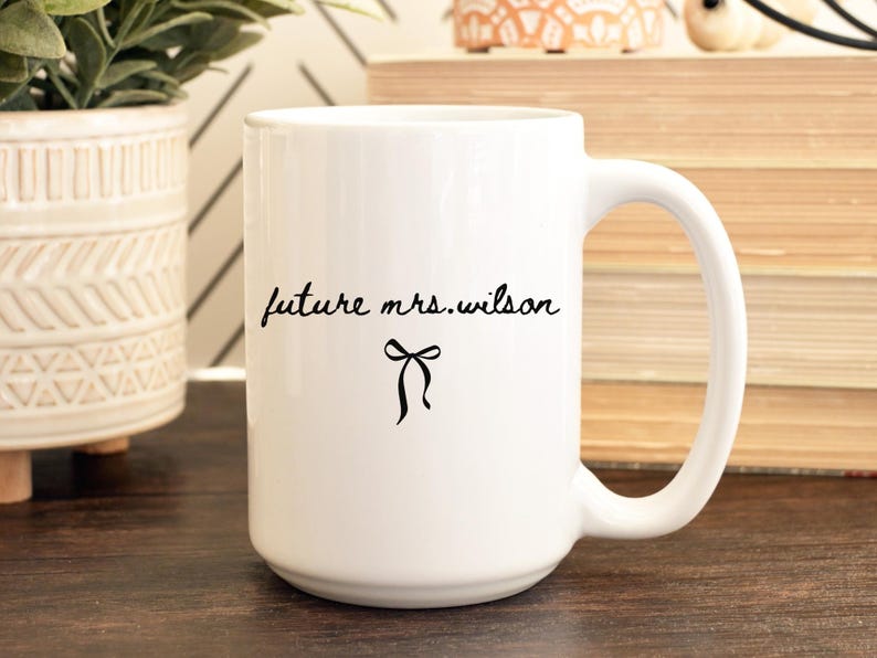 K&ouml;nnte beinhalten: Wei&szlig;er Keramikbecher mit gro&szlig;em Henkel. Der Becher tr&auml;gt den Text "future mrs. wilson" in handschriftlicher Form, mit einer schwarzen Schleifengrafik darunter. Der Becher steht auf einer Holzoberfl&auml;che.