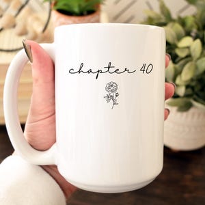 May include: A white ceramic mug featuring the black text "chapter 40" in a cursive script font, positioned above a small, simple black line drawing of a flower. The mug has a smooth, glossy finish and a curved handle on the left side.