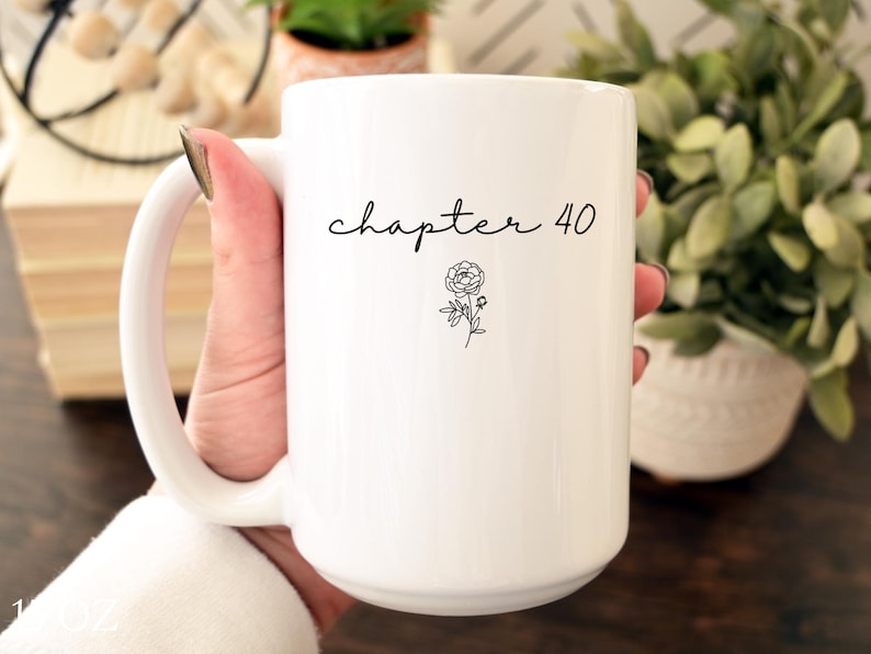 May include: A white ceramic mug featuring the black text "chapter 40" in a cursive script font, positioned above a small, simple black line drawing of a flower. The mug has a smooth, glossy finish and a curved handle on the left side.