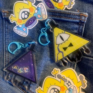 May include: Four colorful stickers featuring a cartoon character with a single eye and a black hole in its stomach. The character is wearing blue sneakers and holding a spoon. The stickers are on a blue denim background. There is also a yellow triangle keychain with a single eye and a black bow tie.