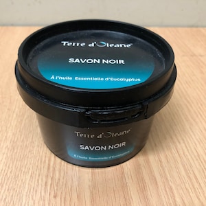 Op de afbeelding: Een zwarte plastic container met een blauw en wit etiket waarop "Terre d'Oleane SAVON NOIR À l'huile Essentielle d'Eucalyptus" staat.