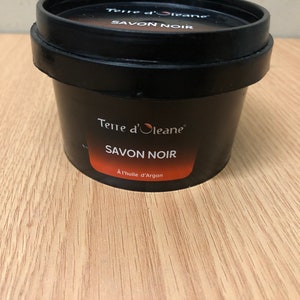 Op de afbeelding: Een zwarte plastic container met Terre d'Oleane Savon Noir zwarte zeep met argan olie. De container heeft een rood etiket met witte tekst.