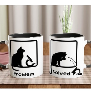 Könnte beinhalten: Zwei weiße Kaffeetassen mit schwarzen Rändern. Die Tasse links hat eine schwarze Silhouette einer Katze, die in ein Mikroskop schaut, mit dem Wort "Problem" darunter gedruckt. Die Tasse rechts hat eine schwarze Silhouette einer Katze, die mit einer Schnur schlägt, mit dem Wort "Gelöst" darunter gedruckt.