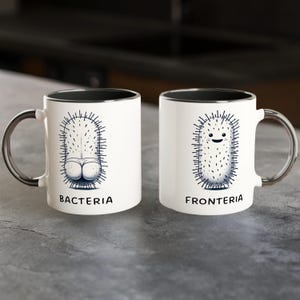Könnte beinhalten: Zwei weiße Keramikbecher mit schwarzen Rändern. Der Becher links hat eine Schwarzweißillustration eines Bakteriums mit dem Wort "BACTERIA" darunter gedruckt. Der Becher rechts hat eine Schwarzweißillustration eines Bakteriums mit einem lächelnden Gesicht und dem Wort "FRONTERIA" darunter gedruckt.