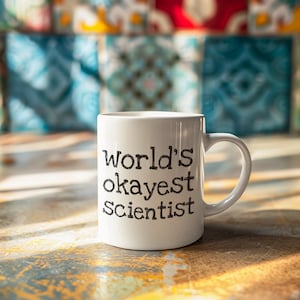 Könnte beinhalten: Eine weiße Keramiktasse mit schwarzem Text, der "World's Okayest Scientist" lautet.