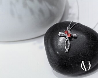 Puede incluir: Un collar con colgante de cruz en tono plateado que presenta dos piedras redondas de color rojo vibrante en el centro. El diseño de la cruz está acentuado con pequeñas piedras claras engastadas en pavé a lo largo del marco metálico curvo.