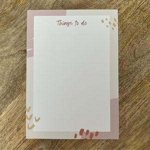 Könnte beinhalten: Rechteckiger Notizblock mit weißem Innenraum und rosa-beigem Rand. Die Worte "Things to do" sind oben in dunkelrosa Schrift gedruckt. Der Rand weist abstrakte Formen in Rosa, Beige und Braun auf.