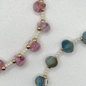 Könnte beinhalten: Nahaufnahme einer Halskette mit rosa und blauen Glasperlen, die durch kleine weiße Perlen und goldene Perlen getrennt sind.
