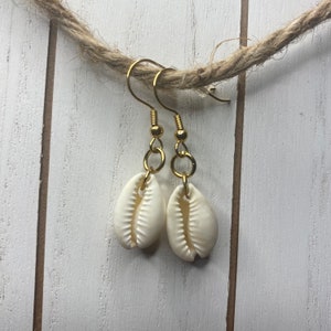 May include: A pair of gold earrings with white cowrie shell charms. The earrings are hanging from a piece of twine.