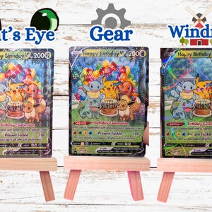May include: Three Happy Birthday V trading cards displayed on small wooden easels. Each card features Pikachu, Squirtle, Charmander, Bulbasaur, and Eevee with a birthday cake and balloons. The cards have distinct holographic patterns: Cat's Eye, Gear, and Windmill.