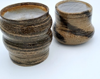May include: Two ceramic cups with brown and white glaze. The cups have a textured, wavy design.