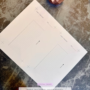 Könnte beinhalten: Zwei weiße Habit-Tracker-Planereinlagen mit Rasterlinien und dem Text "HABIT TRACKER". Die Einlagen liegen auf einer dunkelgrauen Marmoroberfläche. Der Text unten lautet "HABIT TRACKER PLANNER INSERTS Instant Download | Digital File".
