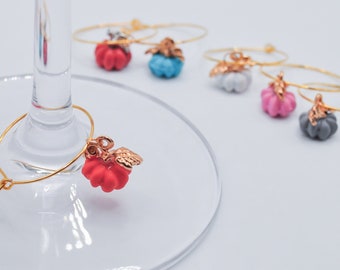 May include: Set of six gold-tone wine glass charms featuring a small pumpkin design in various colors, including red, blue, pink, white, and gray.