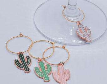 May include: Four gold-colored wine glass charms with cactus designs. The charms are green, pink, and white.