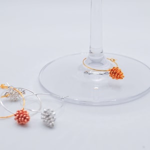 May include: Three gold-tone wine glass charms shaped like pine cones. One charm is on a wine glass, and the other two are on a white surface.