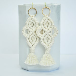 May include: A pair of white macrame earrings with gold hoops and tassels. The earrings are hanging from a white marble surface.
