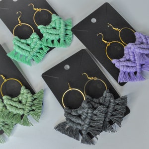 Può includere: Tre paia di orecchini in macramè fatti a mano con cerchi dorati. Gli orecchini sono realizzati con filo verde, viola e grigio e hanno dettagli a frangia.