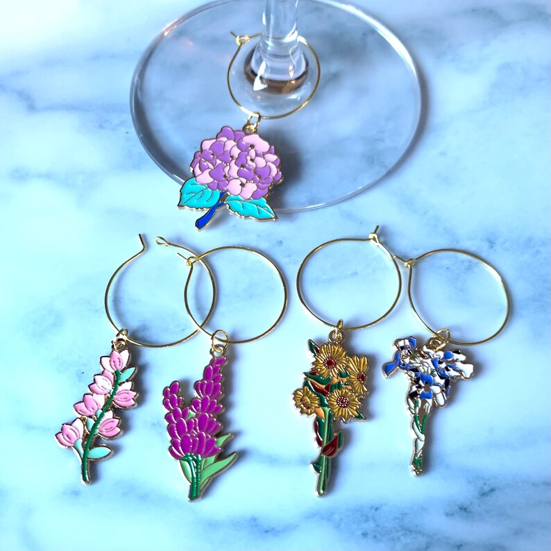 May include: Set of six gold-tone wine glass charms featuring different floral designs. The charms include a pink hydrangea, a pink flower cluster, a purple flower cluster, a yellow sunflower cluster, and a blue and white flower cluster.