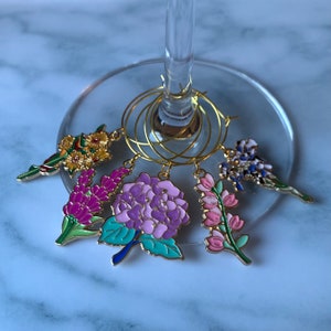 May include: Set of six gold-tone wine glass charms featuring different floral designs. The charms include a purple hydrangea, pink flowers, purple and white flowers, yellow flowers, and blue flowers.