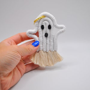 May include: White ghost-shaped macrame wall hanging with a gold halo and black eyes. The ghost has a fringe of beige yarn at the bottom.