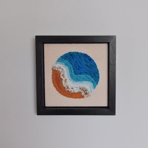 Può includere: Un ricamo incorniciato che rappresenta un'onda oceanica stilizzata con colori blu e bianchi su uno sfondo color sabbia.