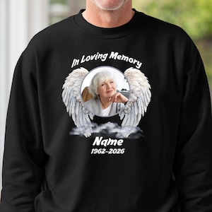 May include: Black sweatshirt with a memorial design. The design features a photo of a person surrounded by angel wings, with the text "In Loving Memory" and the name and dates 1962-2026.
